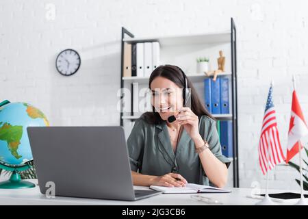 Insegnante positivo in cuffie con microfono che ha lezioni online sul portatile vicino al globo e bandiere a scuola Foto Stock