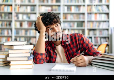 Triste arabo o indiano maschio studente stanco di studiare, si siede a una scrivania in una biblioteca sullo sfondo di librerie, sconvolto ragazzo millenario guarda via, si sente esaurito da lunghi libri di lettura Foto Stock