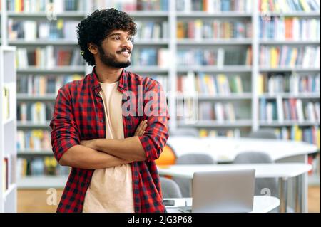 Positivo bello capelli ricci indiano o arabo freelancer o studente, in abiti eleganti moderni, si erge in biblioteca con le braccia incrociate, guarda via, sorridente amichevole, pensiero, sognare Foto Stock