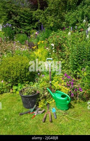 Giardino di campagna inglese all'inizio di luglio. Una selezione di strumenti ben utilizzati accanto ai confini pieni di varie piante. Foto Stock