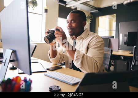 Giovane fotografo professionista nero seduto alla scrivania che lavora su computer Holding Camera editing immagini Foto Stock