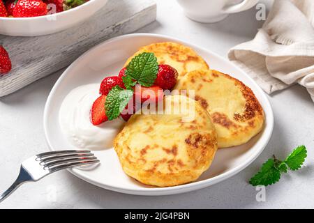Pancake al formaggio casolare con fragole e panna in un piatto bianco su sfondo grigio chiaro da vicino. Foto Stock