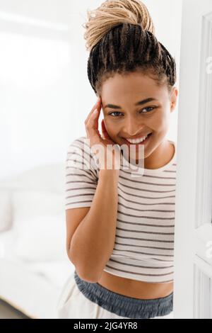 Giovane donna nera con pigtail che sorride mentre si alza in piedi nella porta di casa Foto Stock