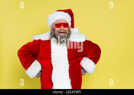 allegro sovrappeso babbo natale in occhiali da sole rossi in piedi akimbo isolato sul giallo Foto Stock