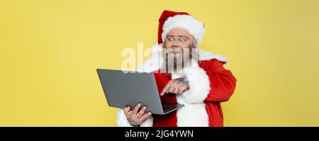 eccitati uomo più formato in costume di natale che punta al computer isolato su giallo, banner Foto Stock