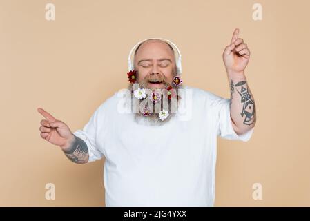 uomo allegro e chubby con musica di ascolto della barba decorata in cuffie isolate sul beige Foto Stock