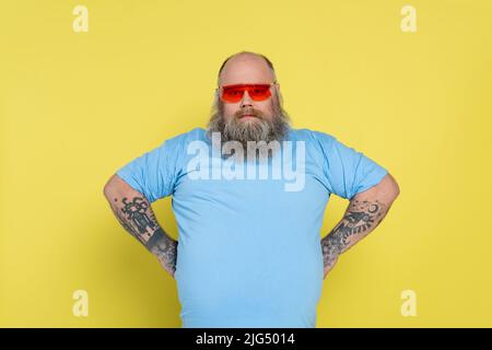 uomo sovrappeso in occhiali da sole rossi in piedi akimbo isolato su giallo Foto Stock