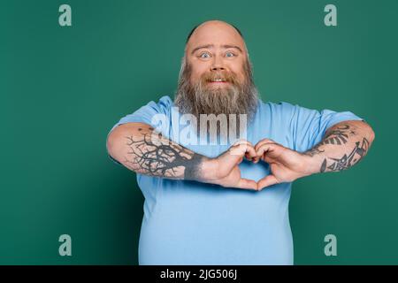 uomo felice più taglia con barba e tatuaggi che mostrano il segno del cuore isolato sul verde Foto Stock