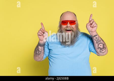 uomo in sovrappeso con bearded e allegro in occhiali da sole che punta in su con le dita isolate sul giallo Foto Stock