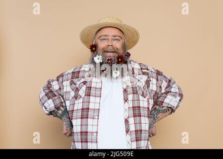 uomo felice in sovrappeso in cappello di paglia e camicia a quadri in posa con fiori in barba isolata sul beige Foto Stock