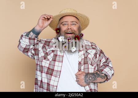 uomo chubby con fiori in barba regolare cappello di paglia e sorridente a macchina fotografica isolato sul beige Foto Stock