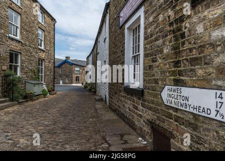 Strada acciottolata e vecchi edifici nel villaggio di Yorkshire Dales con indicazioni per altre città in primo piano che puntano lungo la scena stradale. Foto Stock