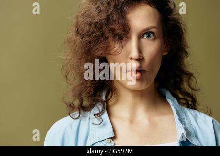 Divertente cute curly bella femmina in jeans casual shirt fa labbra arco come pesce posa isolato su sfondo verde oliva pastello. Essere voi stessi Foto Stock