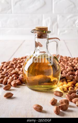 Olio di arachidi in una caraffa di vetro e arachidi crude su un tavolo di legno bianco. Olio da cucina monoinsaturo di Arachis ipogaea. Frutta di arachidi e olio. Foto Stock