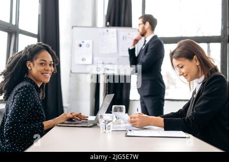 Bella donna d'affari afroamericana di successo, si siede alla scrivania in ufficio, guarda la macchina fotografica, sorridente. Team di lavoratori di uffici multirazziali che lavorano insieme su un progetto in un ufficio moderno Foto Stock