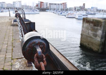 Catena Rusty che fissa un dissuasore in cemento con barche ancorate sullo sfondo in un moderno porticciolo Foto Stock