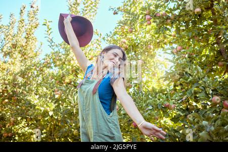 Giovane donna eccitata che salta per gioia in un frutteto di mele in una giornata di sole fuori. Contadino felice e allegro sentirsi ottimista, libero e pieno di energia Foto Stock