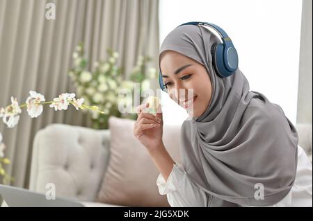 Attraente donna musulmana asiatica millenaria che indossa hijab si rilassa in un comodo soggiorno, mangiando macaron mentre si usa un computer portatile. Foto Stock