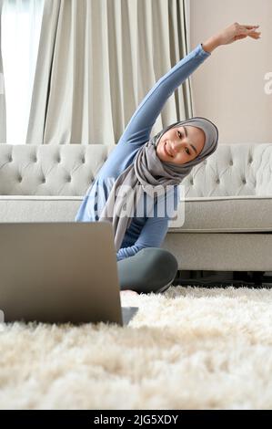 Bella donna musulmana asiatica millenaria che indossa hijab, braccio allungante, yoga imparante pone sul Internet attraverso il suo computer portatile nel vivo r Foto Stock