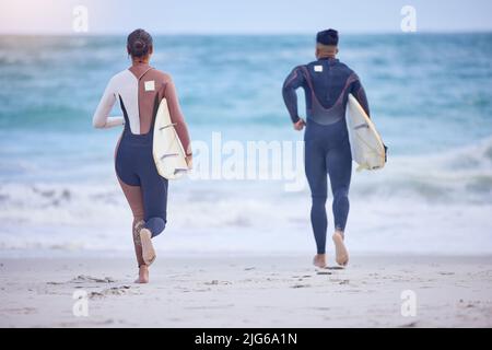 Cavalca come se la possiedi. Ripresa retrostensiva di una giovane coppia che corre verso il mare con le loro tavole da surf. Foto Stock