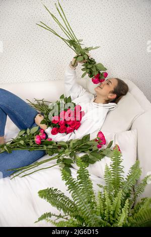 Una ragazza giovane e bella si trova sul letto e felicemente odora rose. Fiori sparsi sul letto Foto Stock