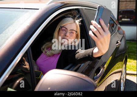 donna bionda di mezza età in occhiali facendo selfie con uno smartphone attraverso un finestrino aperto per auto Foto Stock