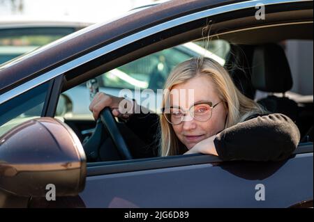 la premurosa bionda di mezza età in occhiali si siede dietro il volante di un'auto, vista attraverso la finestra Foto Stock