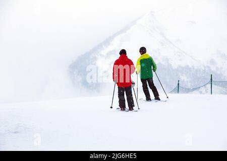 Due sciatori in tute da sci rosse e blu prima di scendere un pendio di montagna sullo sfondo di neve bianca e picchi di montagna in lontananza, Bakuria Foto Stock