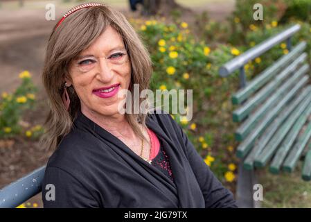 Uomo transgender con make-up su e indossare abiti donna seduti su una panca in un parco pubblico Foto Stock