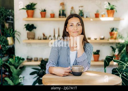 Bella donna brunetta 40s al bar in un negozio accogliente con piante Foto Stock