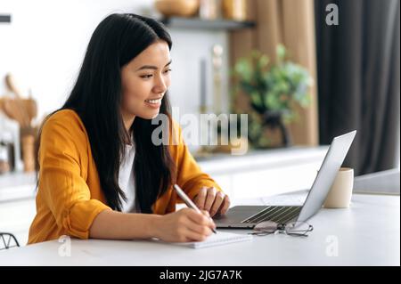 Lezione di distanza, lezione. Fidata ragazza asiatica positiva intelligente, studente di successo, studia da casa utilizzando il laptop, ascolta il webinar online, video lezione, prendere appunti, sorridendo felicemente Foto Stock