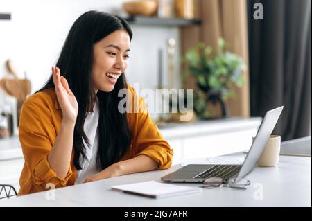 Vista laterale di una ragazza socievole asiatica positiva, in abiti casual, lavorando da casa seduta in cucina, comunica tramite video conferenza online con i dipendenti, saluta, sorriso, discute questioni di lavoro Foto Stock