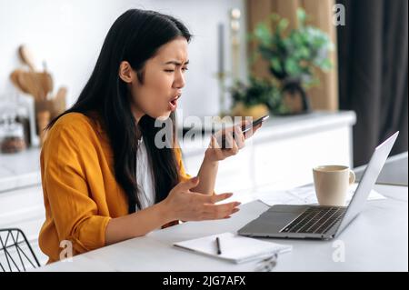 Infastidito giovane donna asiatica, freelance, studente, ceo, si siede a casa in cucina, lavoro a distanza, tenendo in mano lo smartphone parlare in vivavoce, registrare il messaggio audio, espressione emotiva del volto Foto Stock