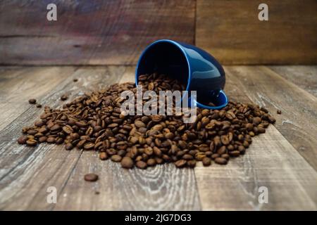 Una tazza di caffè blu viene capovolta con i chicchi che fuoriescono dalla tazza su una superficie di legno. Foto Stock