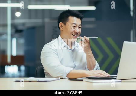Felice imprenditore asiatico che lavora in un ufficio moderno digitando sul portatile, uomo d'affari registrare messaggio vocale, utilizzando app sul telefono per comunicare. Foto Stock