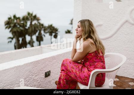 Una donna caucasica in un abito rosa carino seduto su un balcone contro una spiaggia in Messico Foto Stock