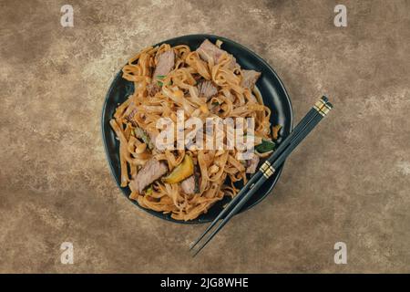 Una ciotola asiatica con bistecca a fette sottili, spaghetti di riso e verdure su un piatto nero con bacchette e sfondo marrone testurizzato. Foto Stock