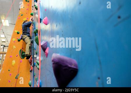 Giovane uomo che si arrampica su una parete coperta di roccia, vista dal basso; corde e sostegni per l'arrampicata per sostenere la salita. Foto Stock