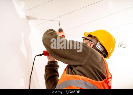 Operatore vestito in uniforme di sicurezza come gilet, casco e occhiali di sicurezza, trapanando un tetto con un trapano. Concetto di lavoro, costruttore, lavoratore. Foto Stock