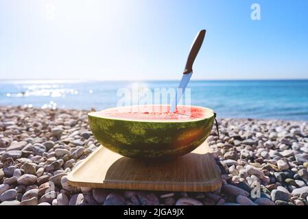 Mezzo anguria sulla spiaggia con ciottoli all'alba. Vacanza in mare con un pic-nic sulla riva. Foto Stock