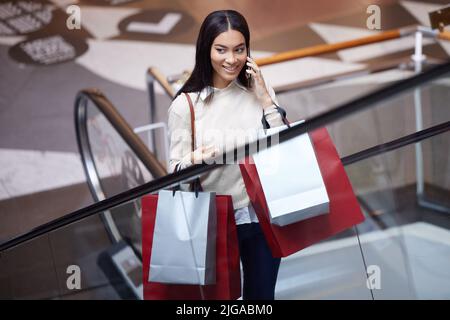 Ho appena finito il mio shopping, voglio prendere un caffè. Una donna che parla sul suo cellulare mentre si va su una scala mobile con borse shopping. Foto Stock