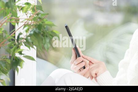 La ragazza si siede accanto alla finestra e usa il telefono cellulare, guarda lo smartphone, legge, acquista, comunica con gli amici in Messenger. Luce brillante immagine Foto Stock