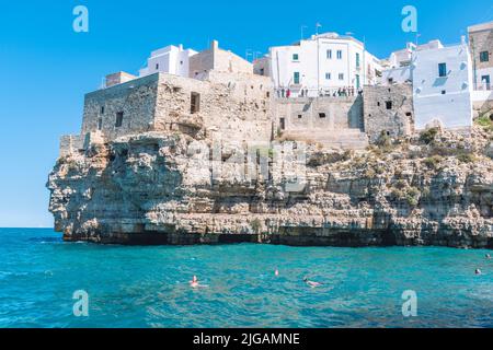 Splendida vista su Polignano a Mare con case in pietra bianca, scogliera, spiaggia, mare blu e turisti, bagnanti, circondati dalla natura mediterranea Foto Stock