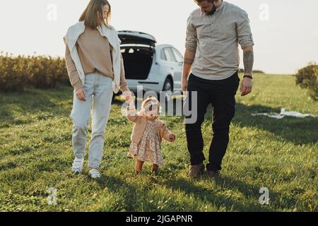 La giovane famiglia si diverta su un'auto SUV, madre e papà tatuato con la bambina che cammina all'aperto sul campo al tramonto Foto Stock