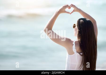 Il mio cuore si trova con l'oceano, una giovane donna che fa un gesto di cuore con le mani in spiaggia. Foto Stock