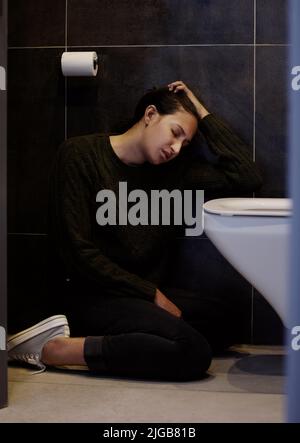 Mi sento orribile. Scatto a tutta lunghezza di una giovane donna attraente seduta accanto al gabinetto nella sua stanza da bagno e sensazione di malessere. Foto Stock
