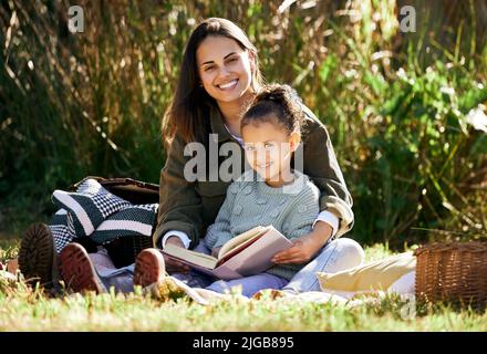 Una bambina e sua madre che leggono un libro mentre si rilassano in un parco o in un giardino. Giovane donna con un bambino piccolo che impara ed ottiene un'istruzione Foto Stock