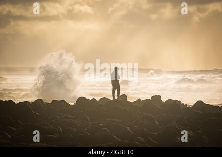 uomo in piedi sulla roccia sulla costa delle isole canarie di lanzarote durante il tramonto con alte onde che si infrangono contro le pietre Foto Stock