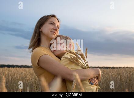 il ragazzo triste abbraccia la madre, si alza su un campo di grano, una donna tiene un bambino tra le braccia e spikelets di grano. Famiglia, rifugiati, unità, sostegno, n Foto Stock