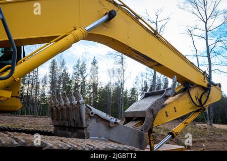 Primo piano di un braccio usato, di una benna e di un cilindro idraulico di una pala caricatrice gommata SSL gialla, in un cantiere edile, alla luce della sera con alberi Foto Stock